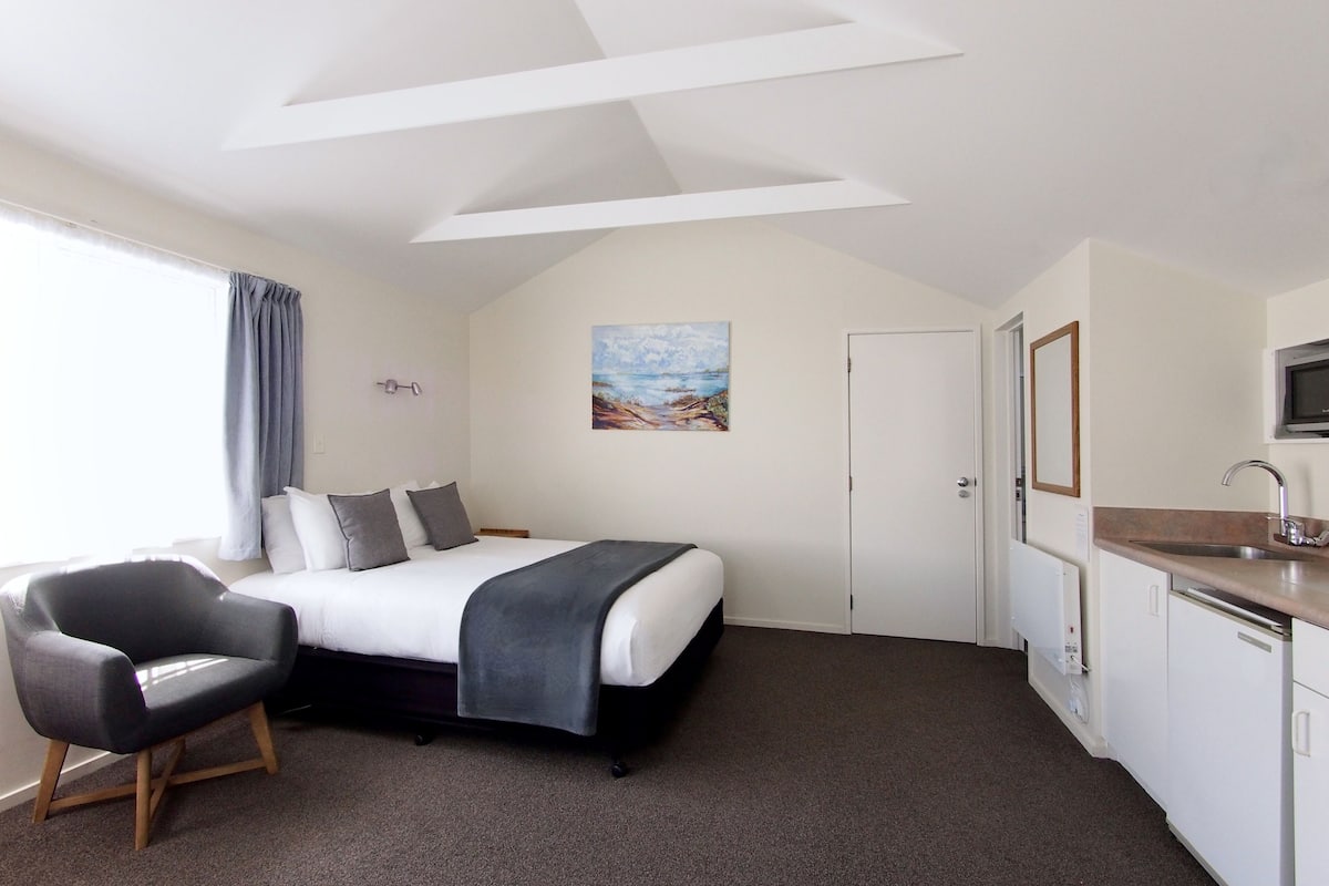 A bedroom with a bed, a chair, a painting, a door, and a kitchenette.