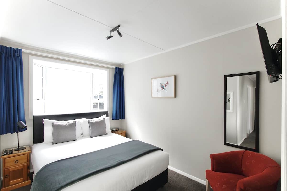 A hotel room with a bed, a chair, a bedside table, a lamp, a window with curtains, a framed picture, and a mirror.