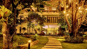 Une entrée d’hôtel bien éclairée, avec un chemin menant au bâtiment principal, entourée d’une végétation luxuriante et de jeux de lumière décoratifs.
