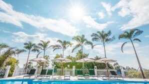 Un espace piscine agrémenté de chaises longues et de parasols, entouré de palmiers sous un ciel dégagé.