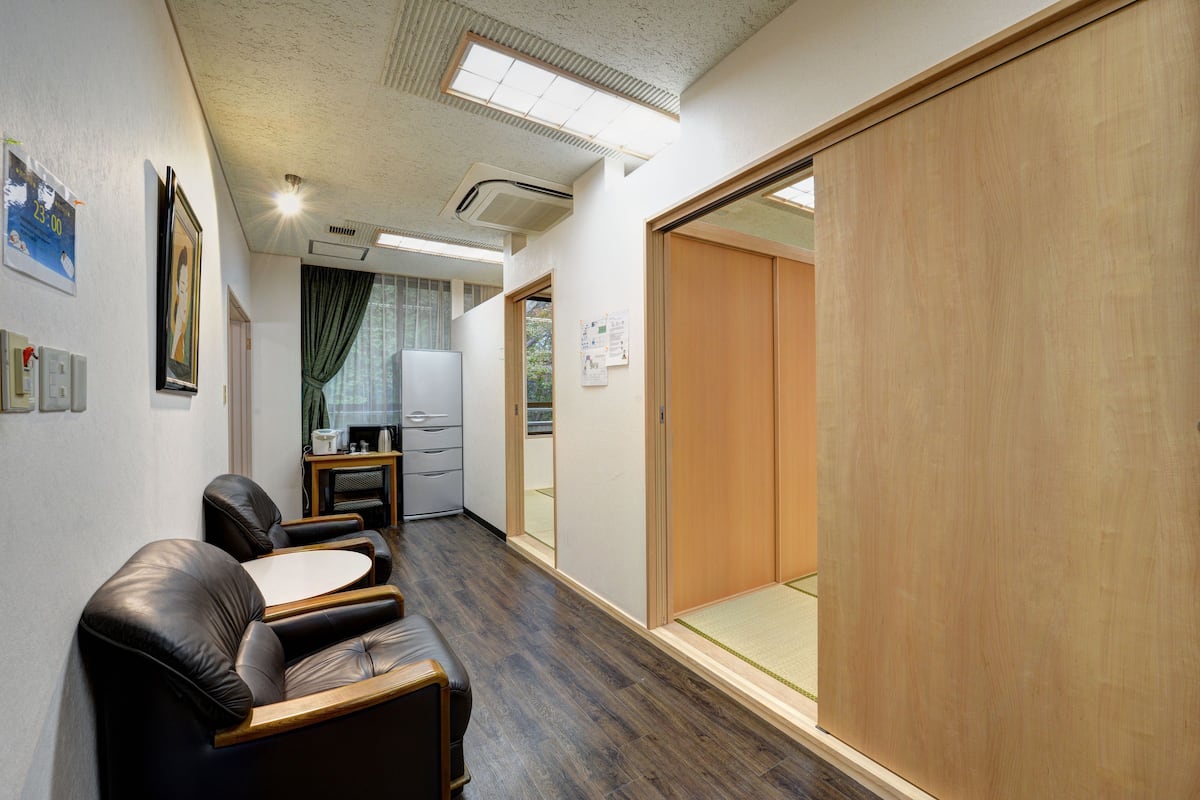 A room with a wooden wardrobe, a sofa, a small table, and a desk with a chair. There is a window with curtains and a door leading to another room.