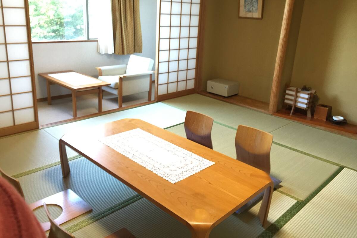 A traditional Japanese room with tatami flooring, a low wooden table, two chairs, a small table, a sliding door, and a futon bed.