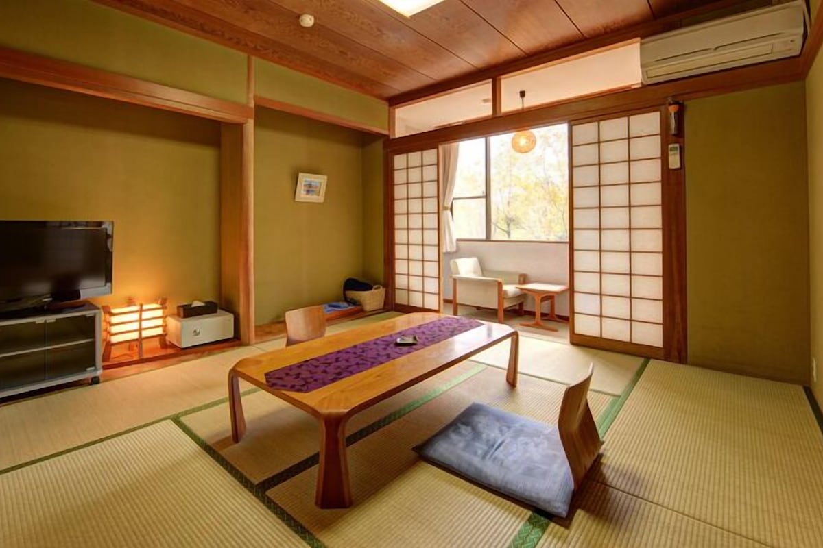 Una habitación tradicional japonesa con suelo de tatami, una mesa baja, un televisor y puertas correderas.
