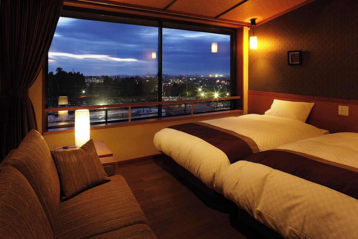 A hotel room with a large window, two beds, a sofa, and a nightstand with a lamp.