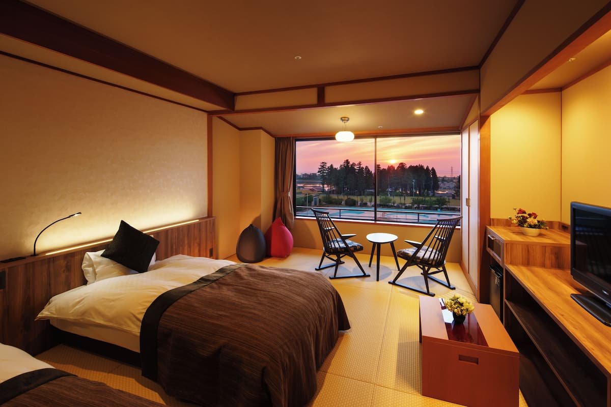 A hotel room with a bed, a desk, a chair, a TV, and a view of the sunset through the window.