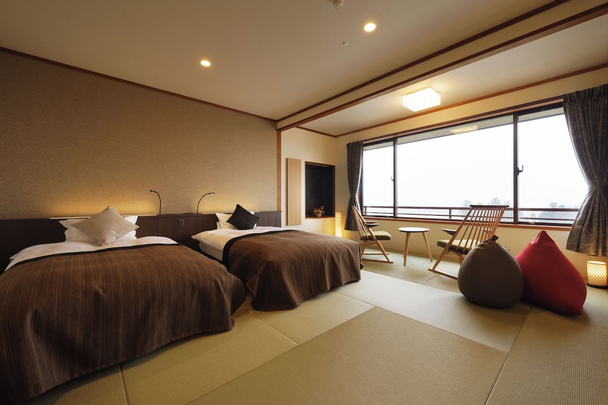 A hotel room with two beds, a sitting area with a chair and a table, and a large window offering a view.