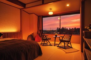 A hotel room with a bed, two rocking chairs, a small table with a vase, and a large window with a view of the city skyline at sunset. - Kintarouonsen (Uozu)