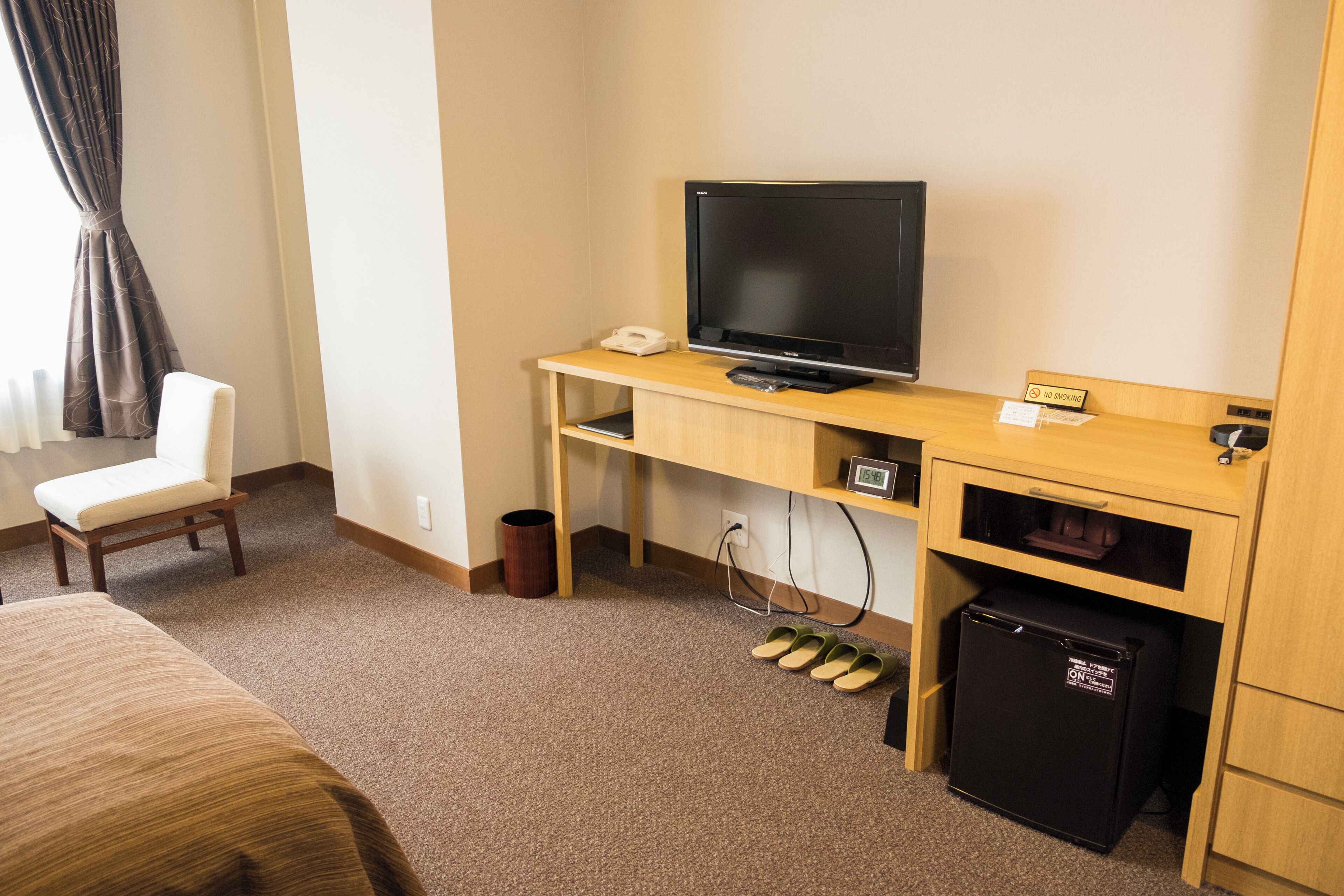 A hotel room with a bed, a desk with a television, a chair, a refrigerator, and a pair of slippers.