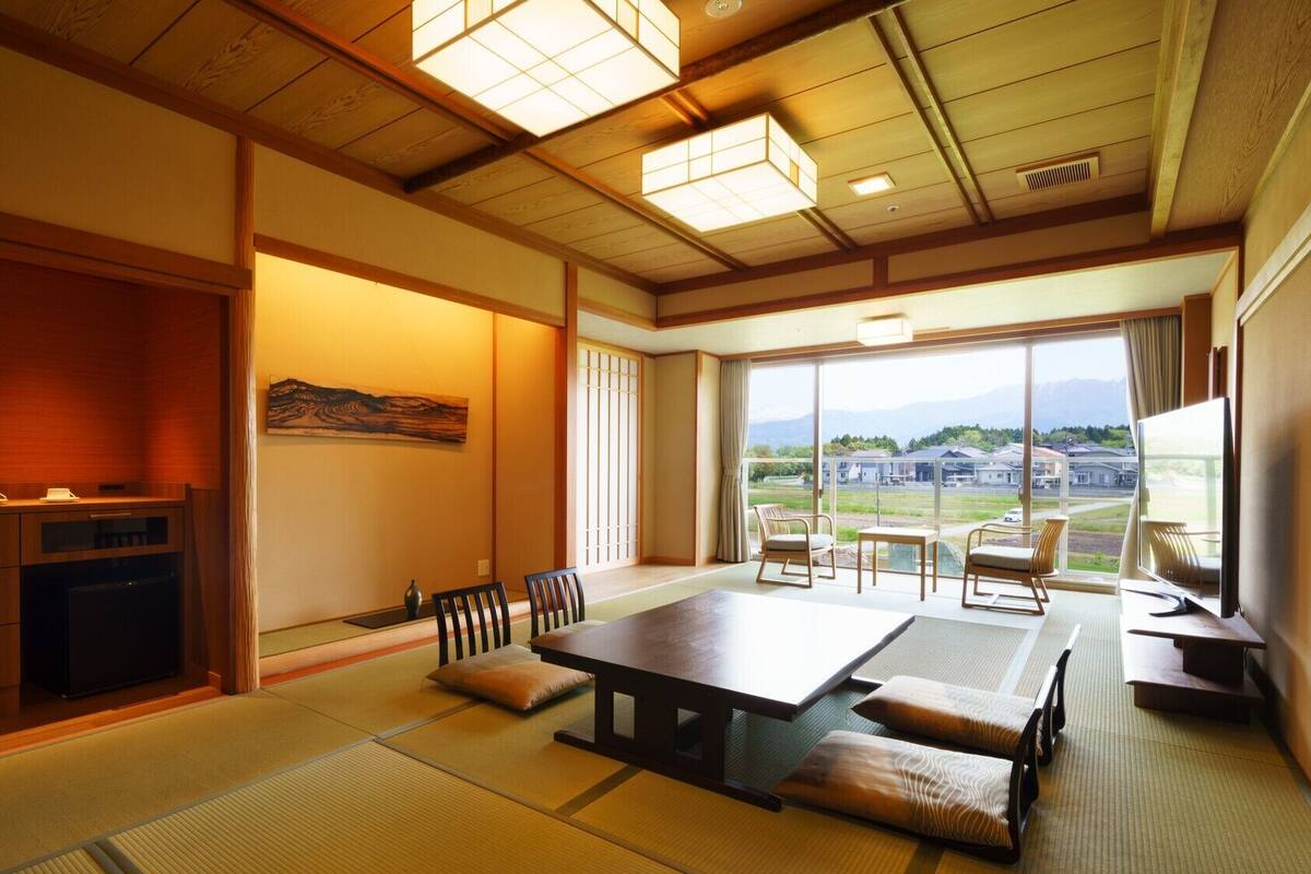 A traditional Japanese room with tatami flooring, a low table, chairs, a television, and a view of a residential area.