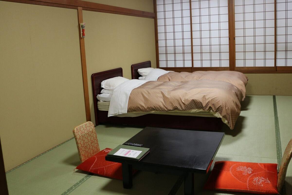 A traditional Japanese room with tatami flooring, a bed with a wooden headboard, a small table with a book, and a futon-style sofa.