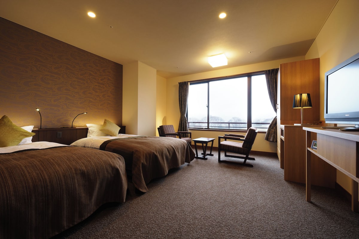 A hotel room with two beds, a desk, a chair, a TV, and a window with a view.
