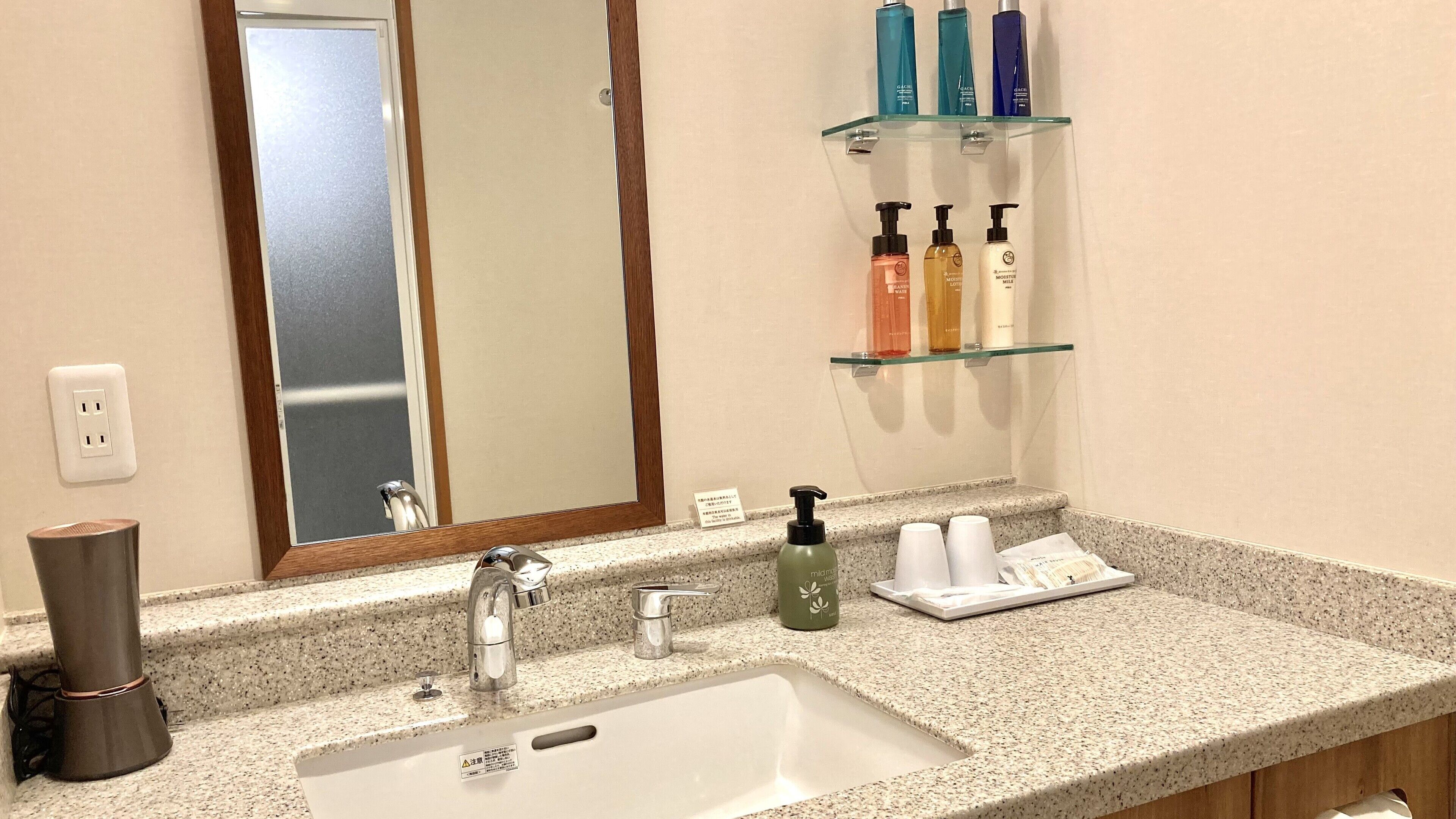 A bathroom with a granite countertop, a white sink, a mirror, and shelves with toiletries.