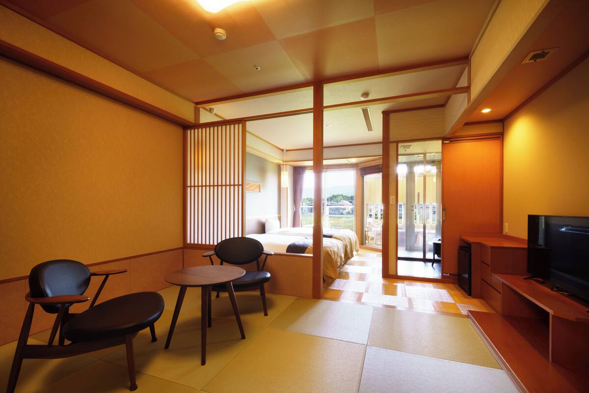 A hotel room with a bed, a TV, a seating area with chairs and a table, and a view through a sliding door to the outside.