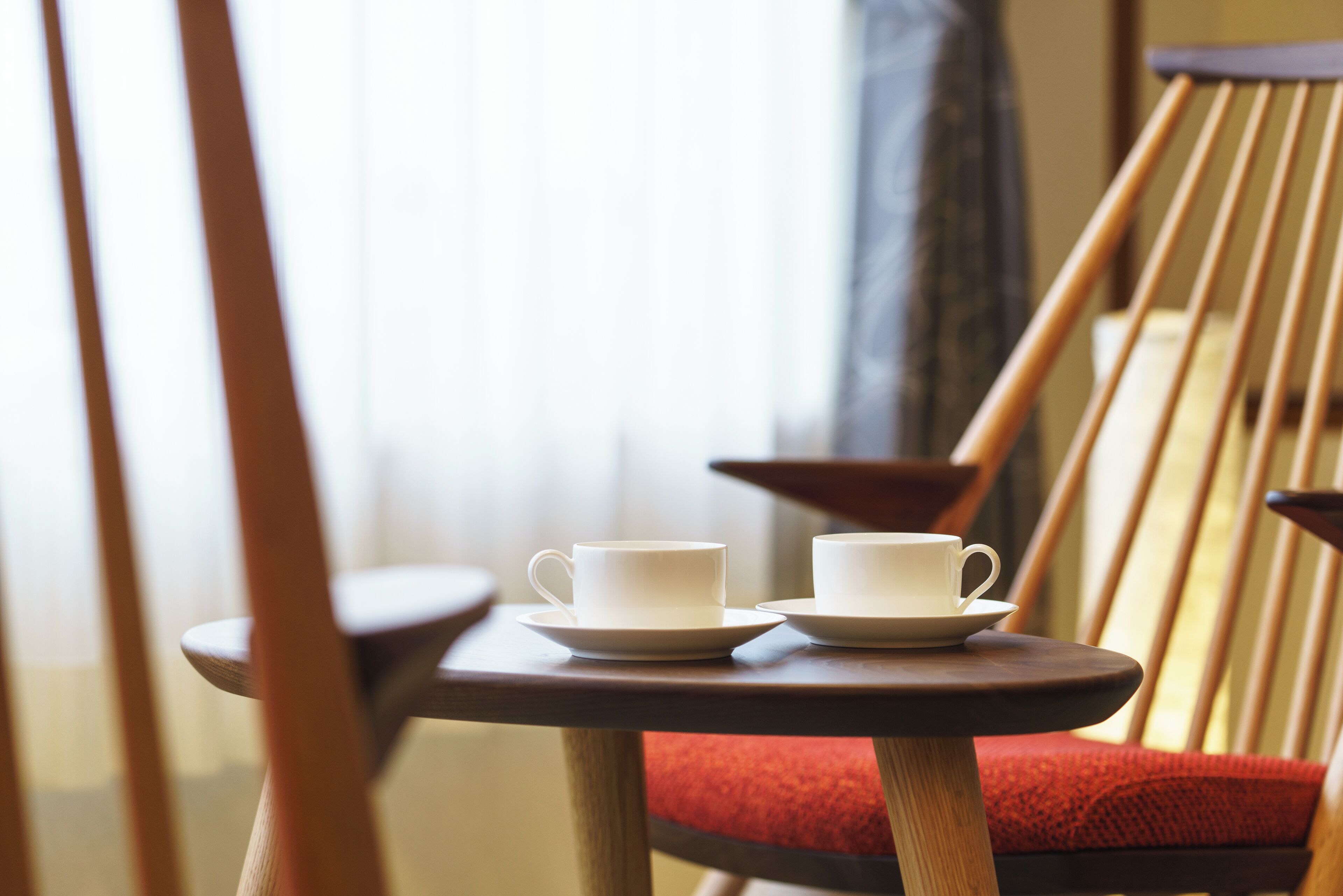 Una silla de madera con cojín rojo y una mesita redonda pequeña con dos tazas y platillos blancos.