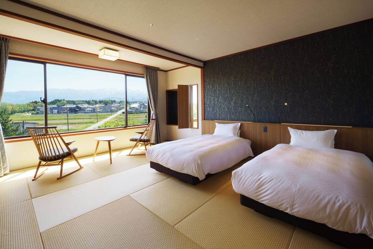 A hotel room with two beds, a view of a mountainous landscape, a wooden rocking chair, and a small table.