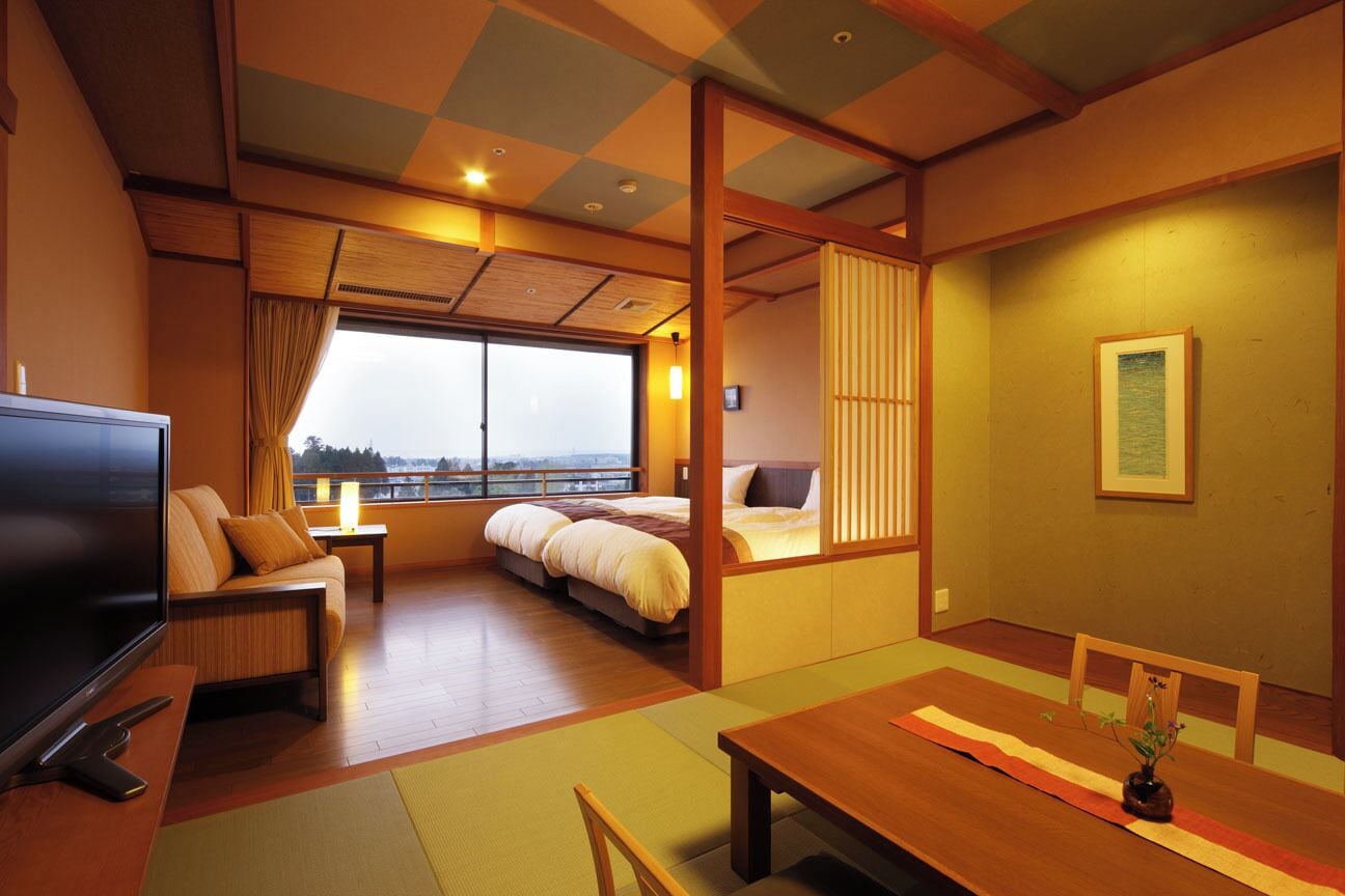 A traditional Japanese room with tatami flooring, a bed, a TV, and a dining table with a view of the city.
