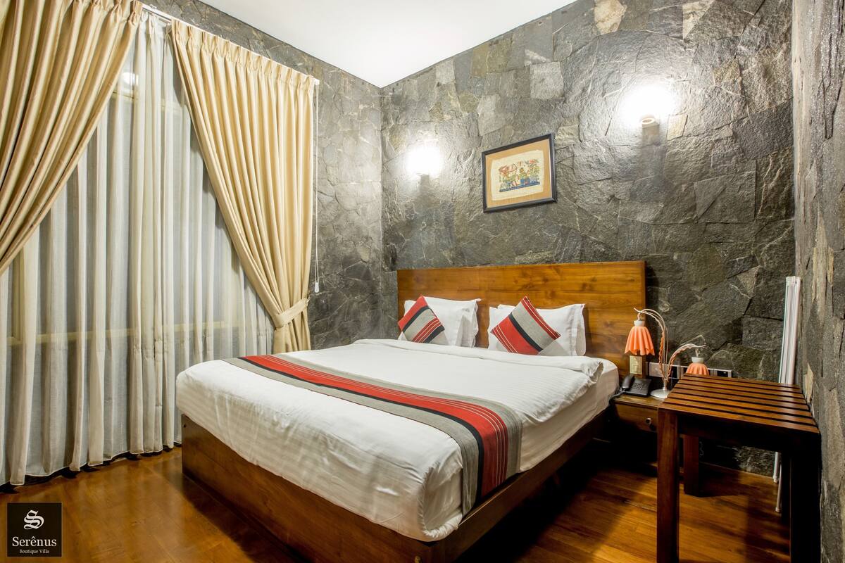 A hotel room with a wooden bed, curtains, a stone wall, and a nightstand with a lamp.