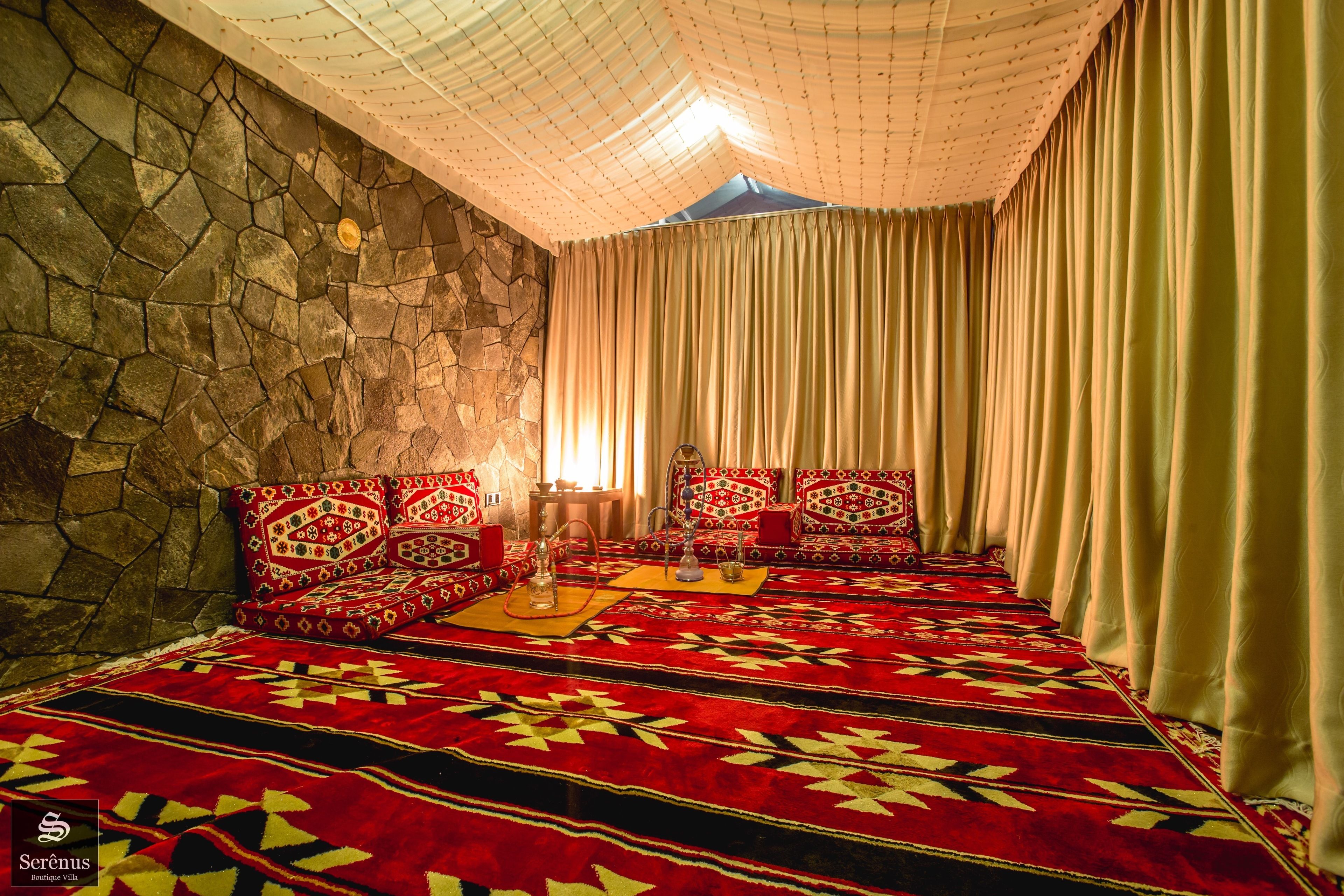 A room with a stone wall, a red patterned carpet, a sofa set with cushions, and a large window with curtains.