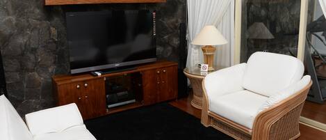 Una sala de estar moderna con un televisor de pantalla plana, un sofá de mimbre, un sillón blanco y un mueble de entretenimiento de madera.