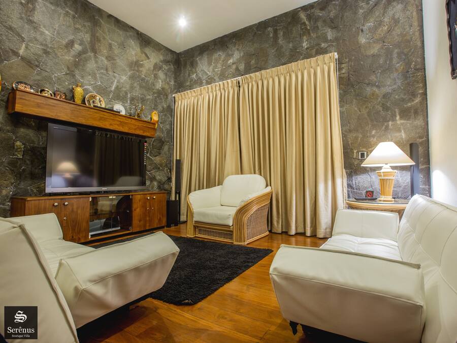 Een woonkamer met een stenen muur, een flatscreen televisie, een witte bank, een rieten fauteuil en een groot raam met beige gordijnen.