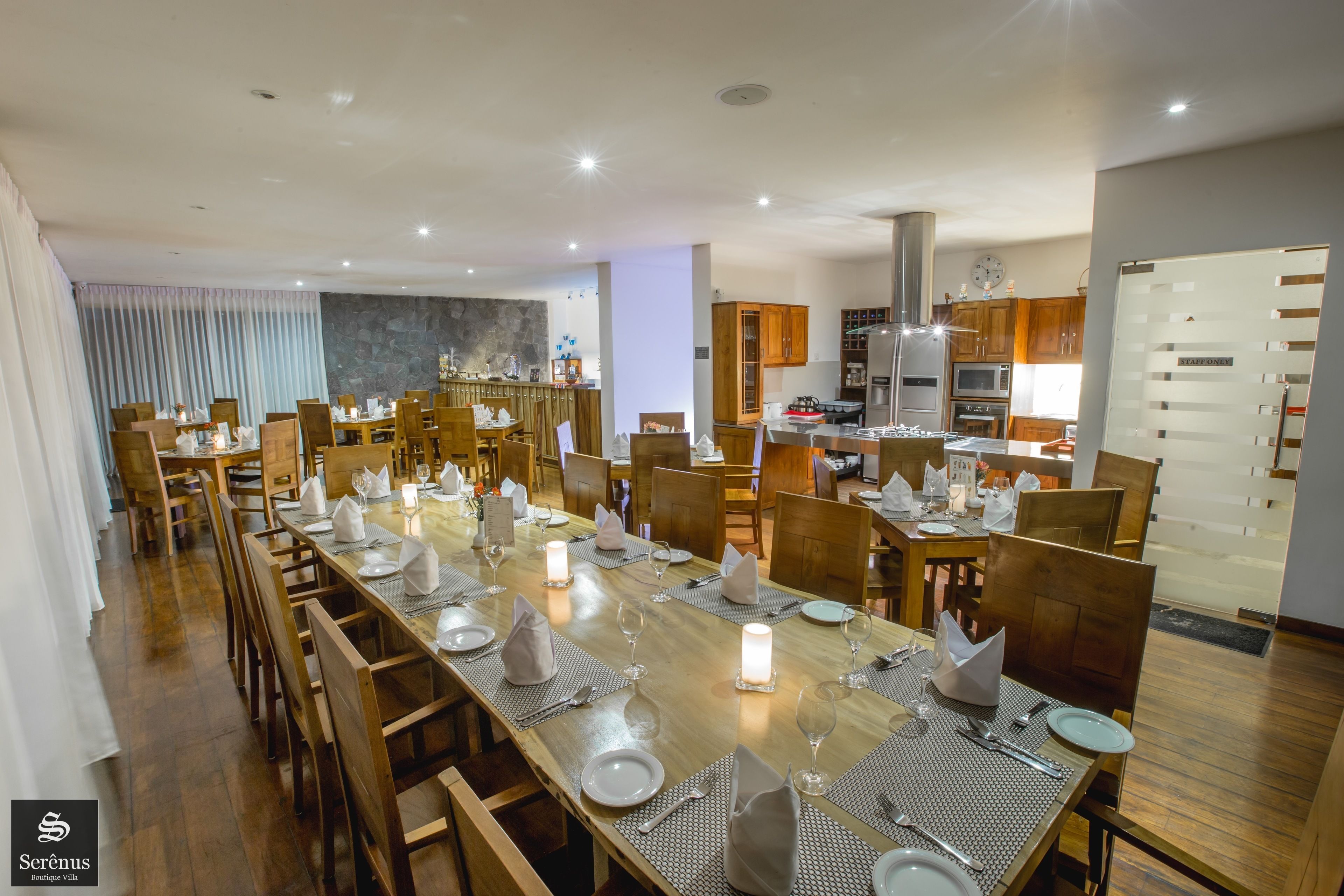 A spacious dining area with multiple tables set for guests, wooden chairs, and a modern kitchen in the background.