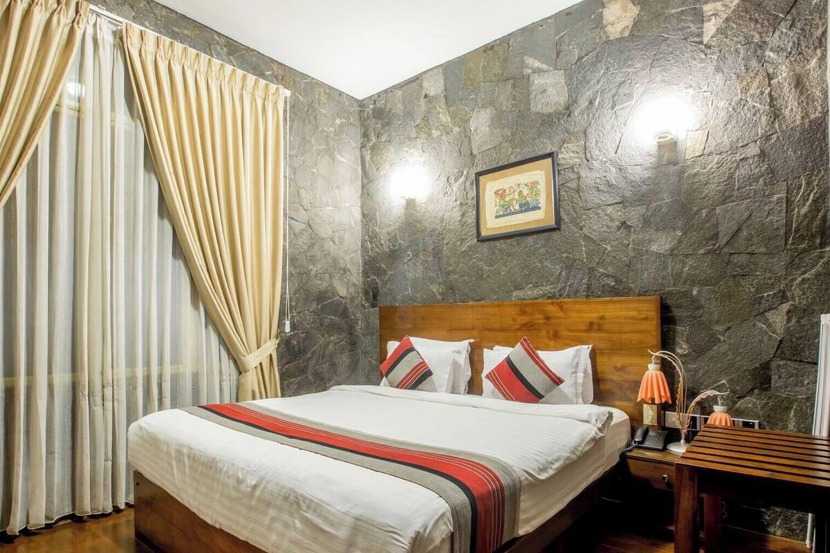 A hotel room with a bed, curtains, a nightstand, and a stone wall.