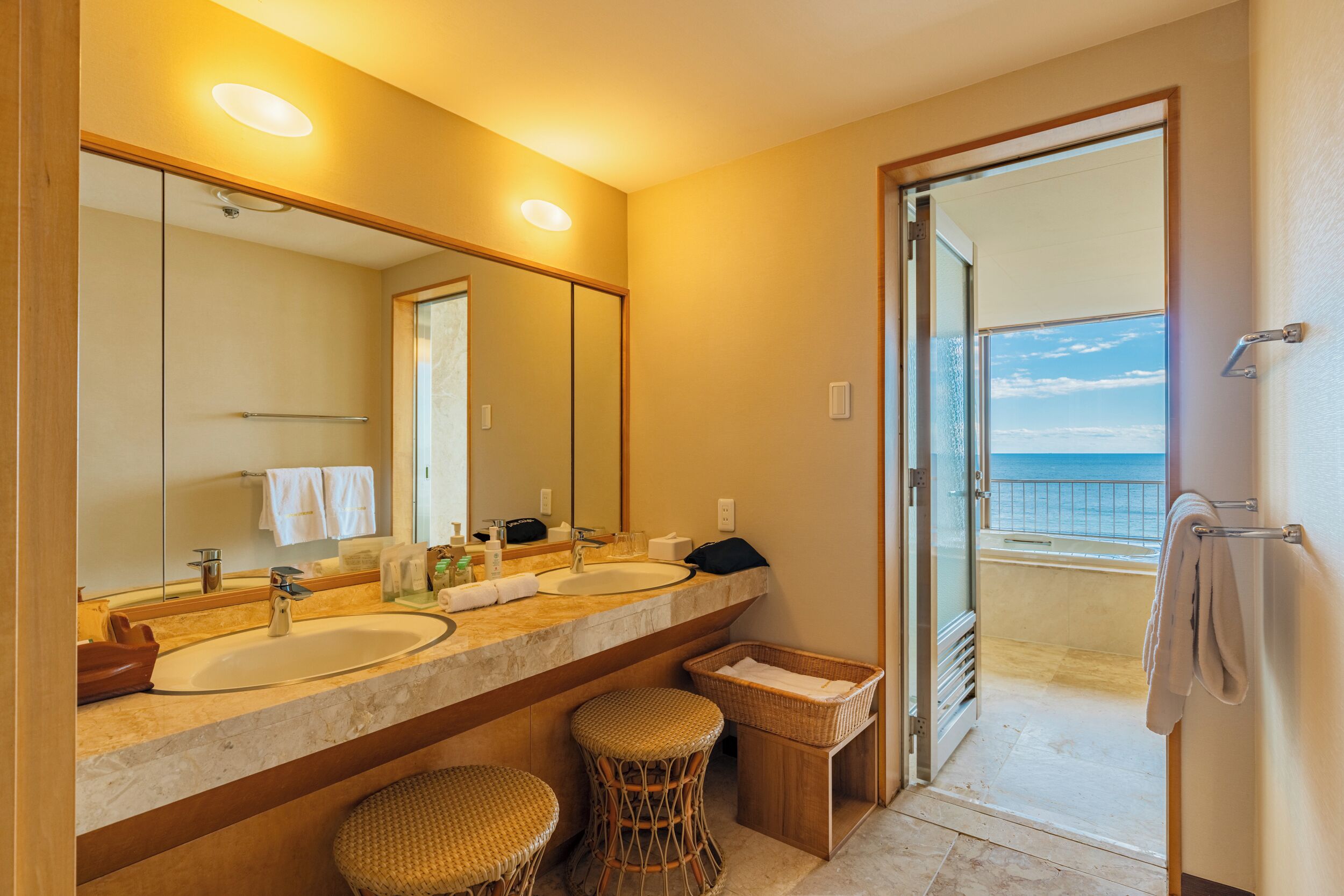 Un baño con dos lavamanos, un espejo grande y vista al mar a través de una puerta abierta.