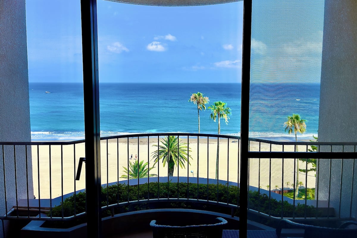 Un balcon avec vue sur l’océan et la plage, agrémenté de palmiers et offrant un ciel dégagé.