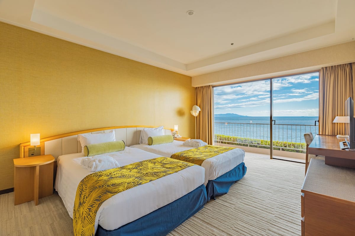 Een hotelkamer met twee bedden, een bureau, een stoel, een lamp en een groot raam met uitzicht op zee.
