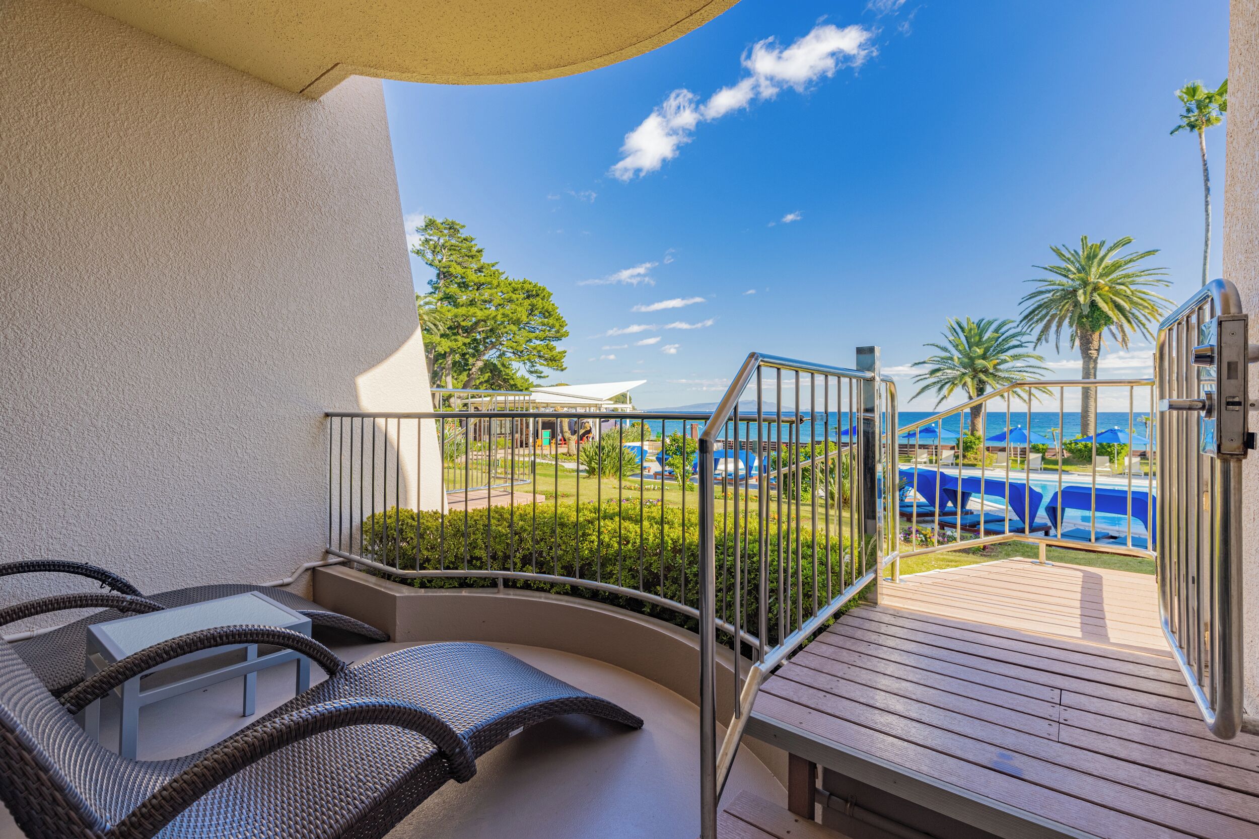 A balcony with wicker furniture, a pool with umbrellas, and palm trees.