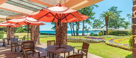 An outdoor patio with wooden tables and chairs, red umbrellas, and a view of the ocean.