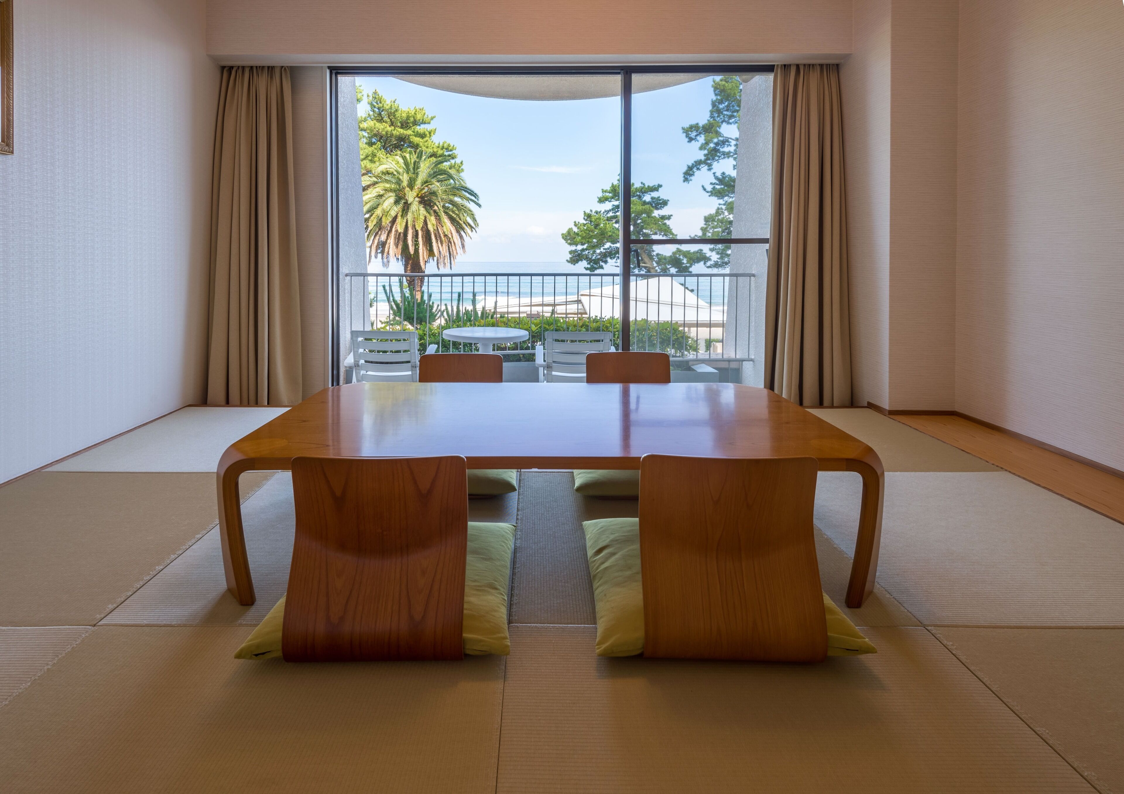 A room with a wooden table and chairs, a sliding glass door leading to a balcony with a view of the sea and palm trees.