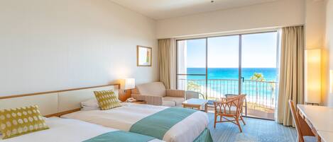 A hotel room with two beds, a balcony with a view of the ocean, a desk, and a chair.