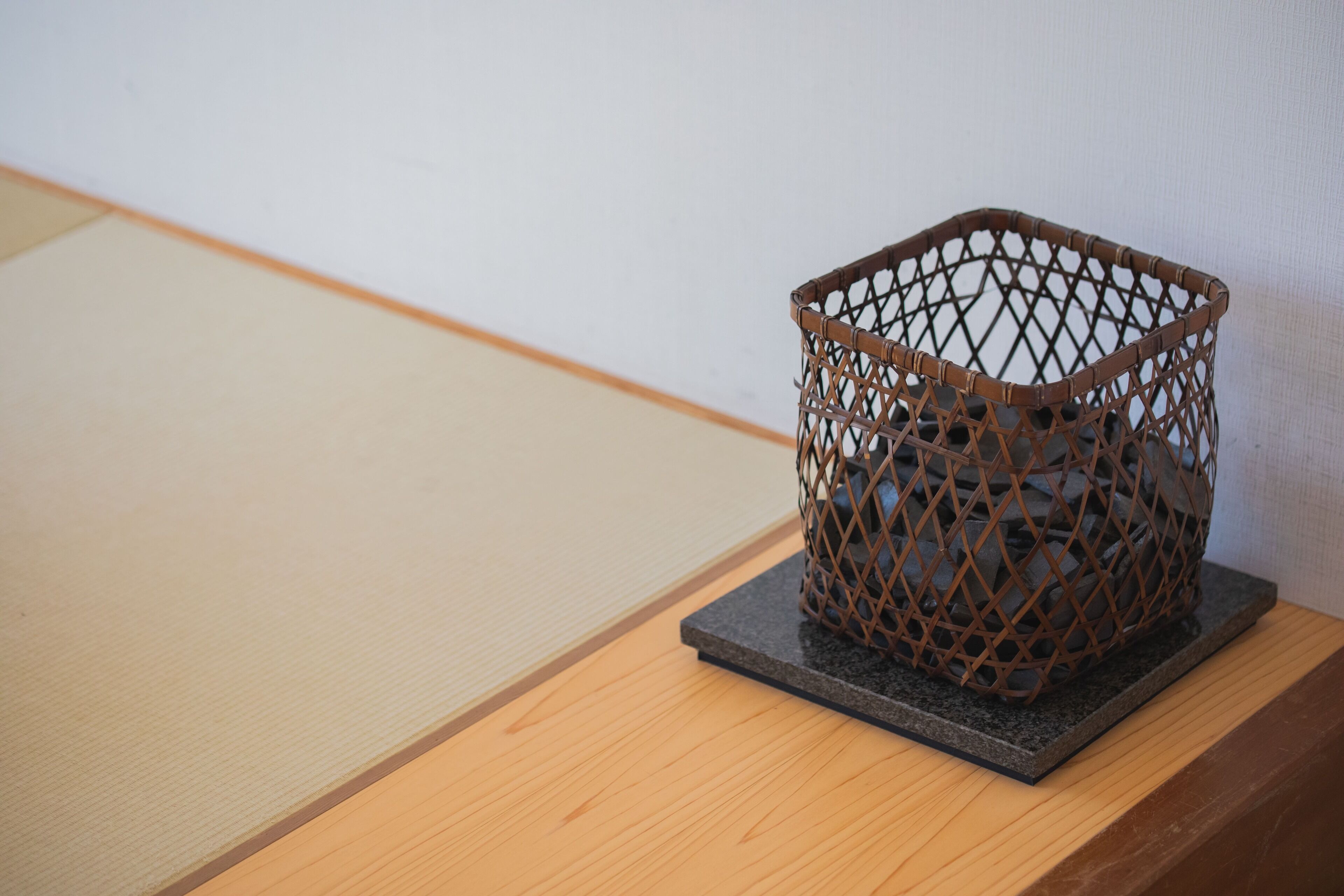Una cesta tejida sobre una base cuadrada oscura, colocada sobre una superficie de madera con un esterillón de tatami al fondo.