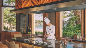 A chef preparing food at a counter with a view of the beach and trees.