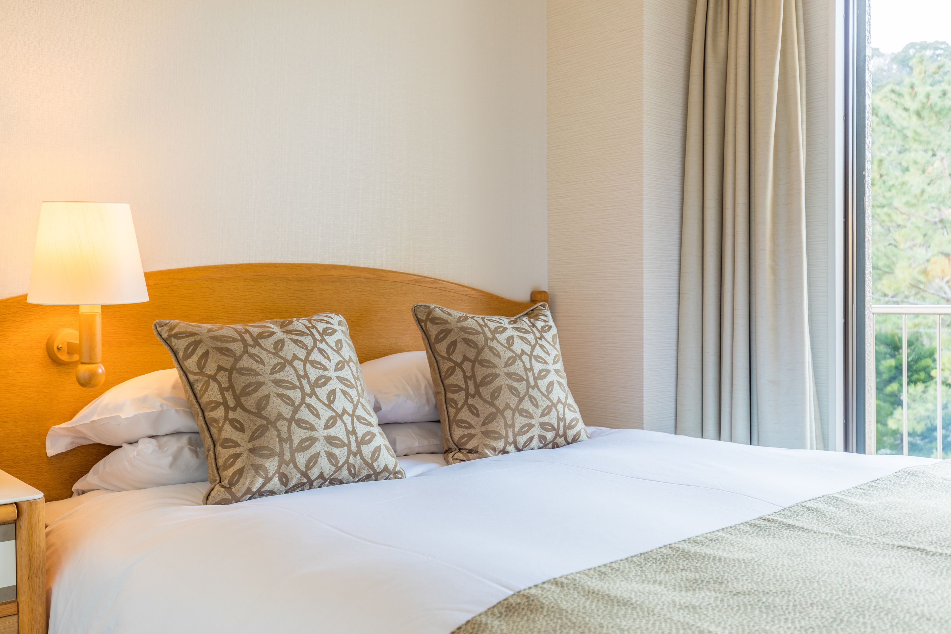 Una cama bien hecha con almohadones estampados, una lámpara de noche y vistas al verdor a través de una ventana.