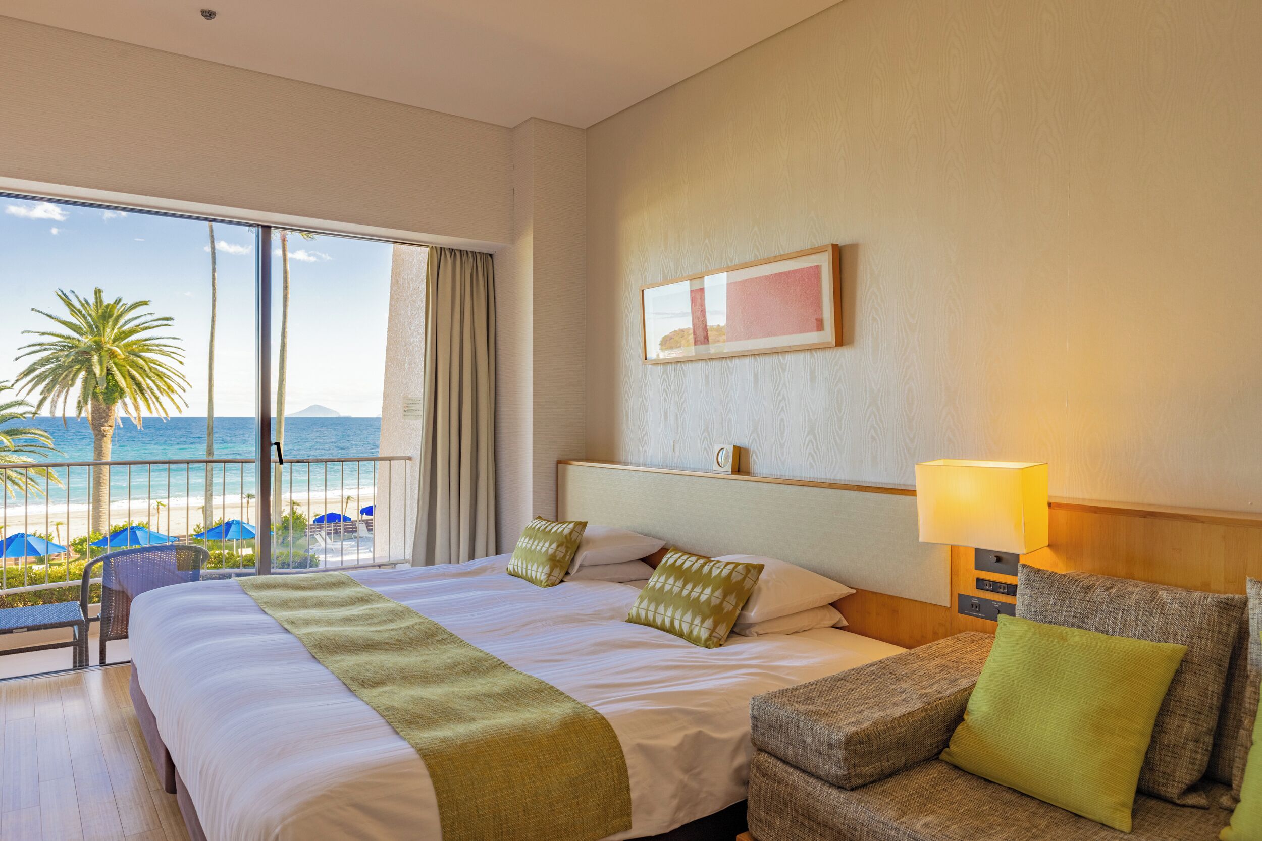 A hotel room with a large bed, a bedside table with a lamp, a view of the beach, and a window with curtains.