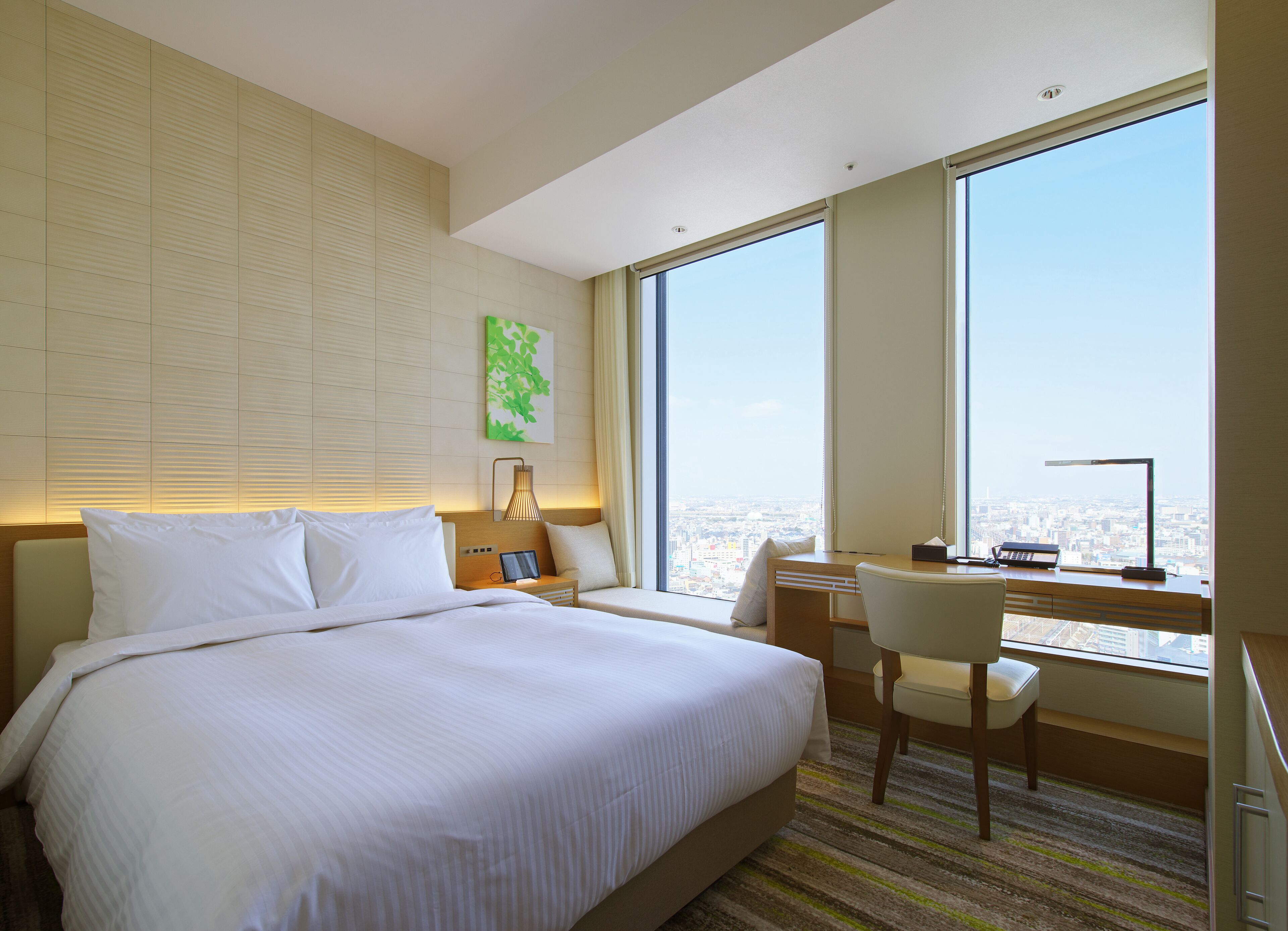 A hotel room with a large bed, a desk with a chair, a view of the city through large windows, and a painting on the wall.