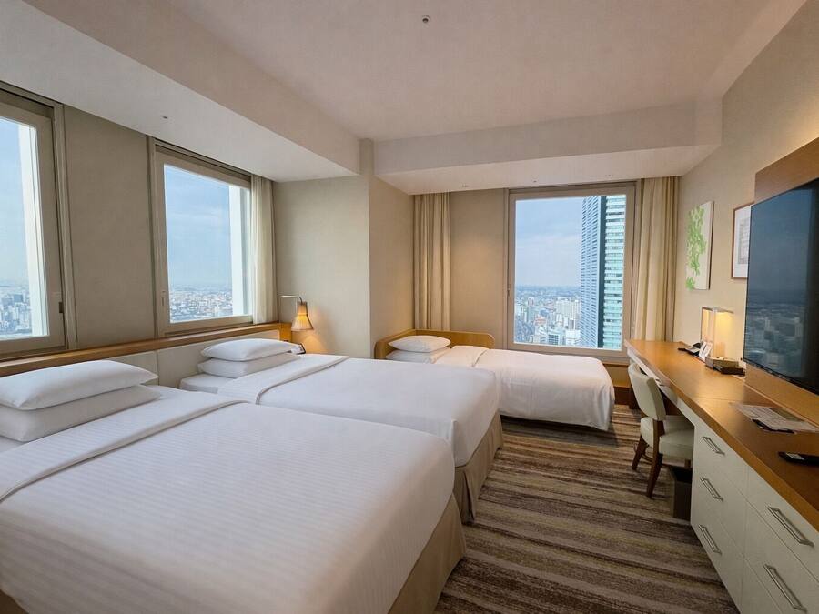 A hotel room with two beds, a desk, a TV, and large windows offering a city view.