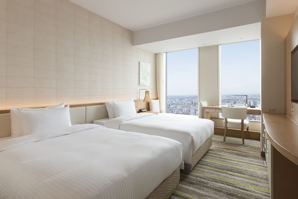 Hotelkamer met twee bedden, een bureau, een stoel en een groot raam met uitzicht op de stad.