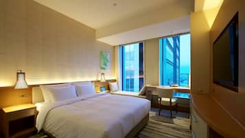 A hotel room with a large bed, a desk, a chair, a TV, and a window with a view of a building.