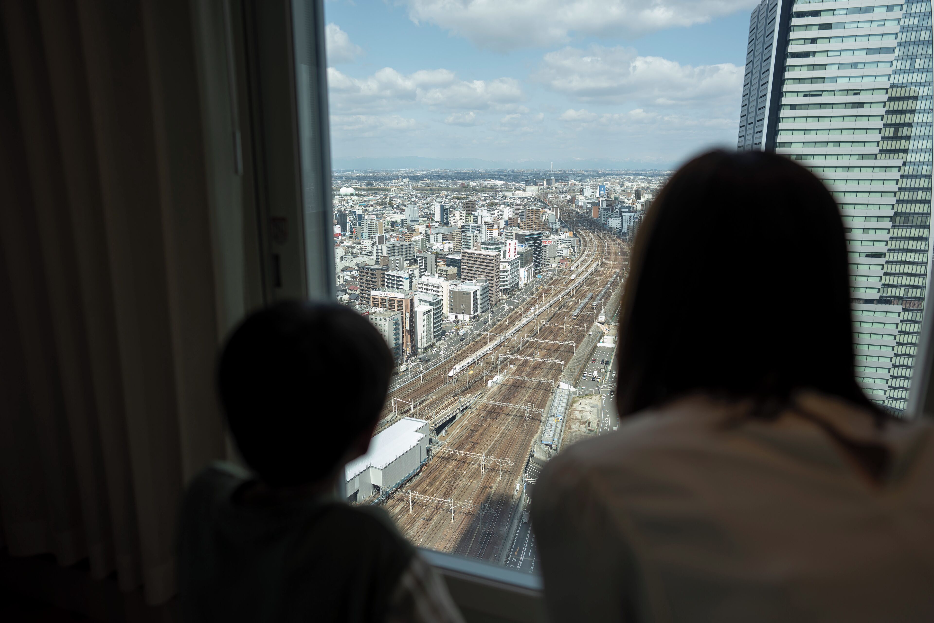 Due persone che guardano attraverso una finestra su un panorama urbano con edifici e binari ferroviari.