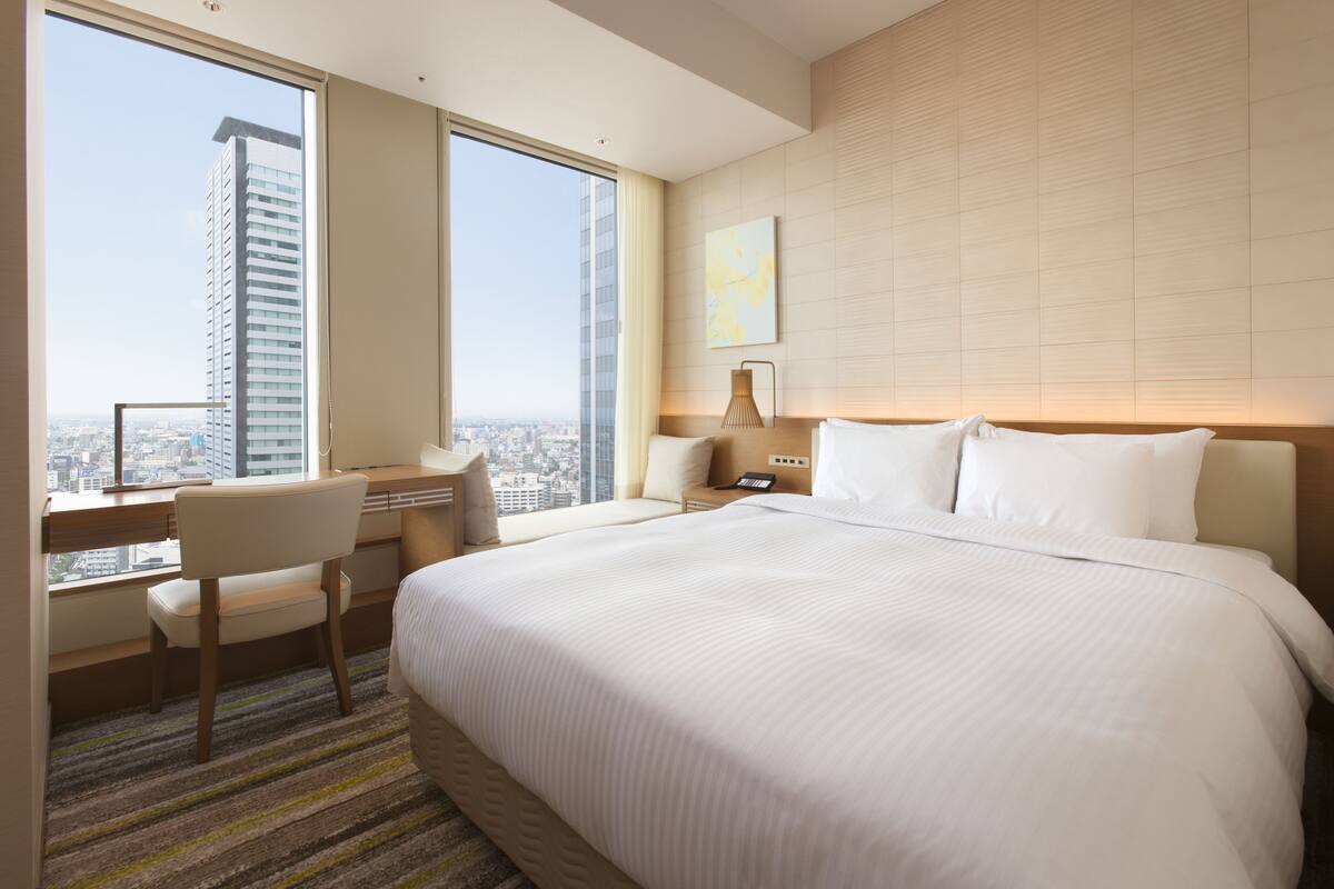 Een hotelkamer met een groot bed, een bureau met stoel, een nachtkastje en een raam met uitzicht op de stad.