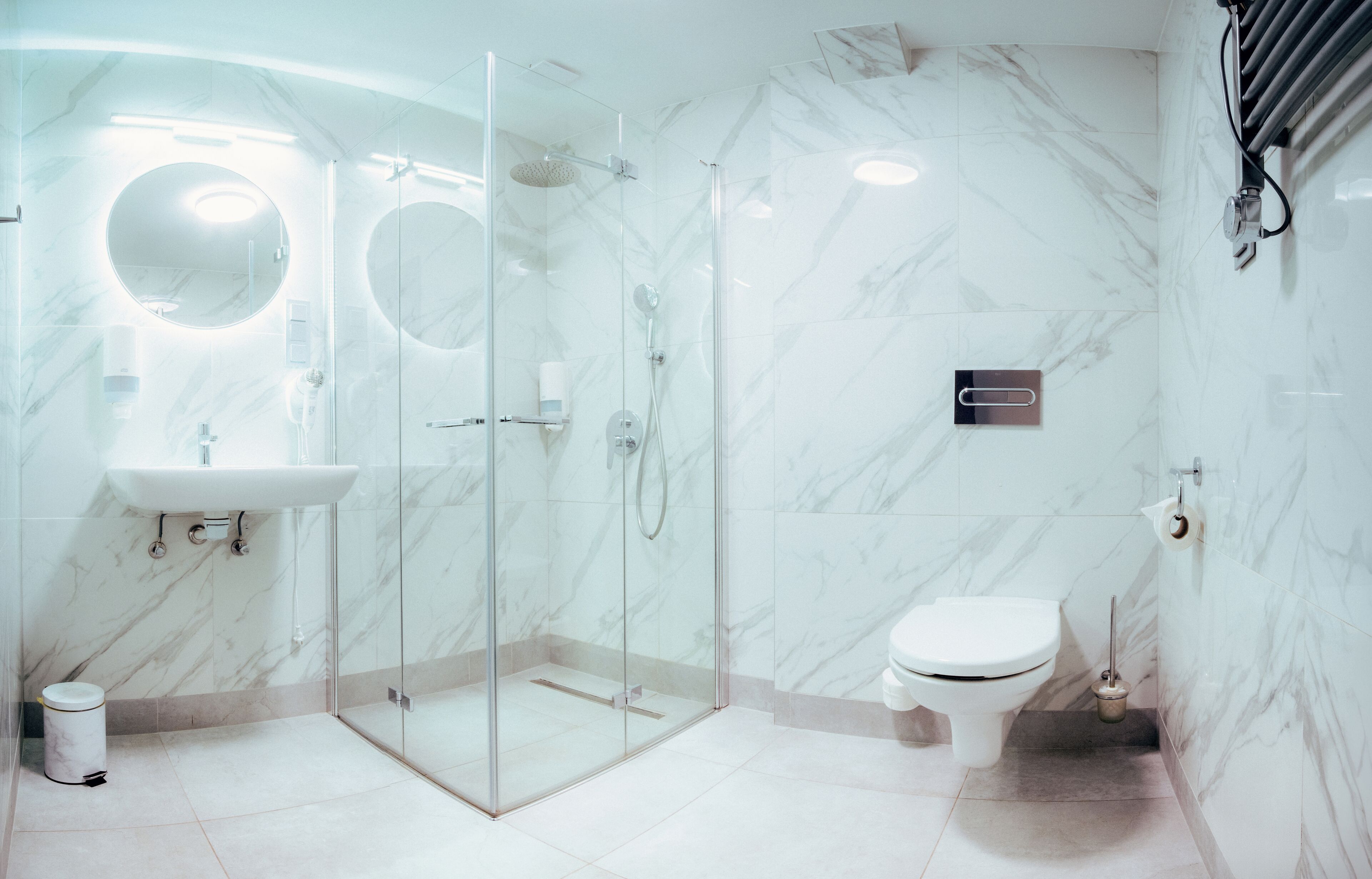 A modern bathroom with a glass-enclosed shower, a sink, two round mirrors, a toilet, and a marble-tiled wall.