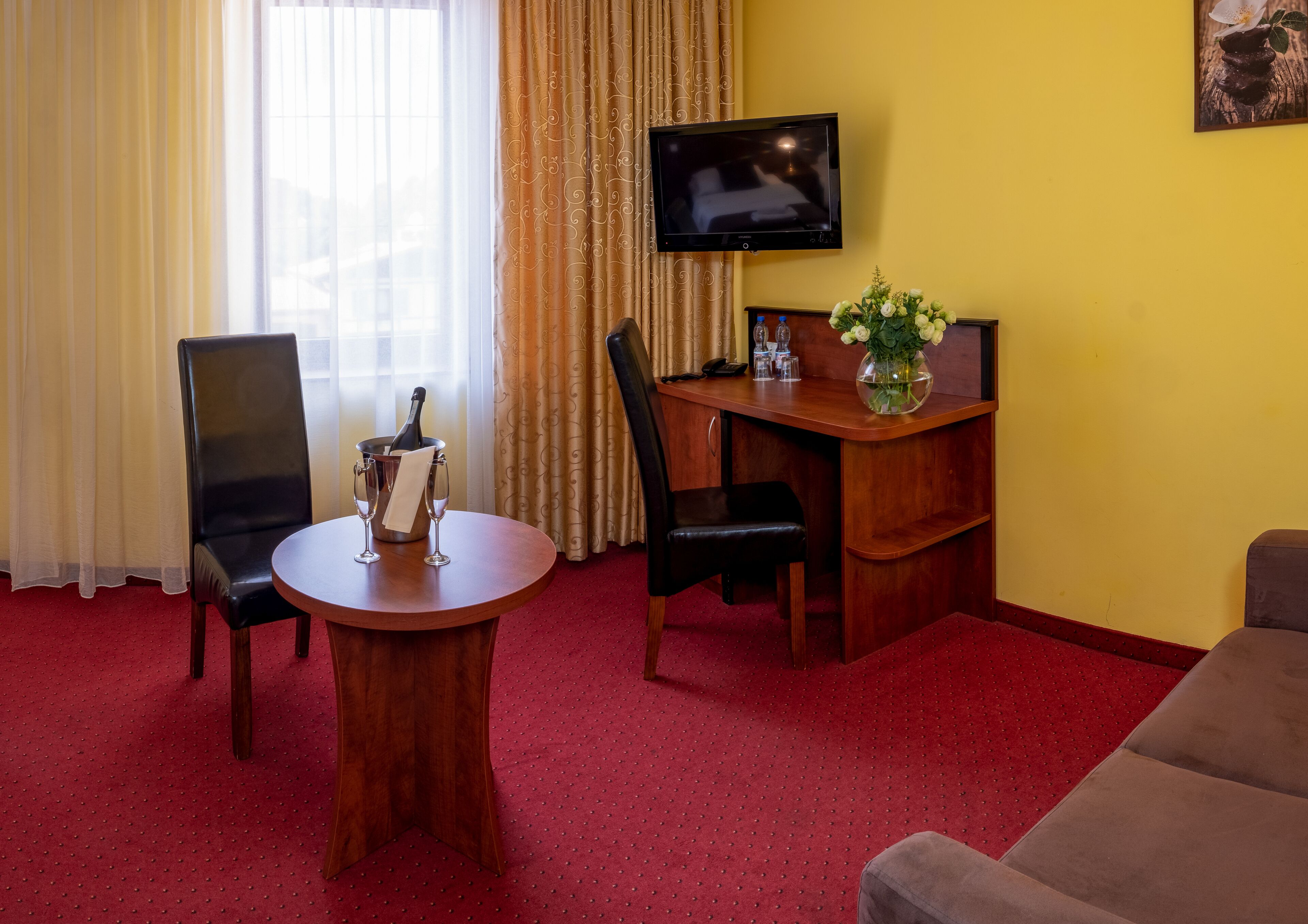 A hotel room with a wooden desk, two chairs, a television, a vase with flowers, and a red carpeted floor.