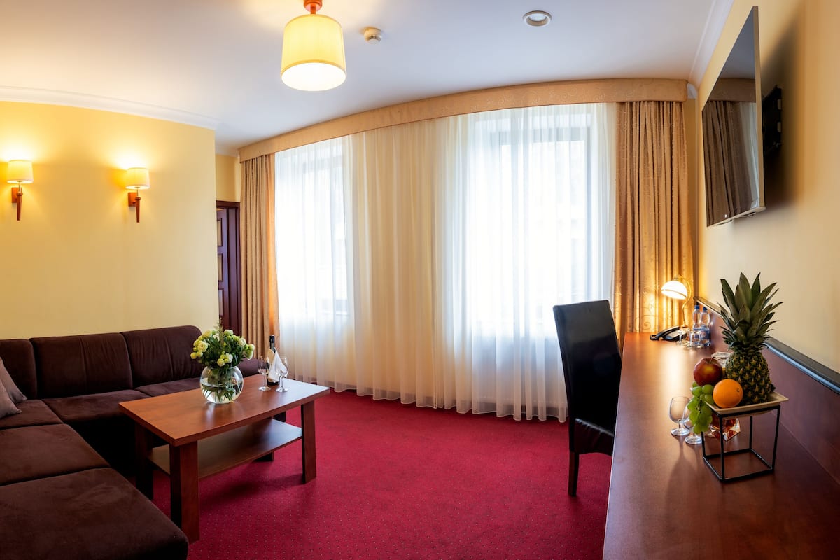 Une chambre d’hôtel avec un canapé marron, une table basse en bois sur laquelle est posé un vase de fleurs, une petite table ronde avec un ananas, une pomme et des raisins, et un tapis rouge.