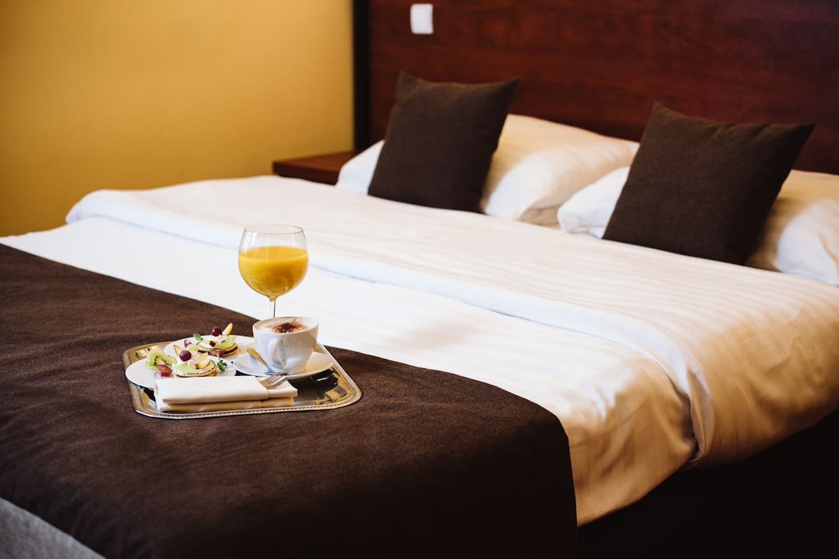 Un lit bien fait, avec un plateau de petit-déjeuner comprenant des toasts, des fruits et un verre de jus d’orange.
