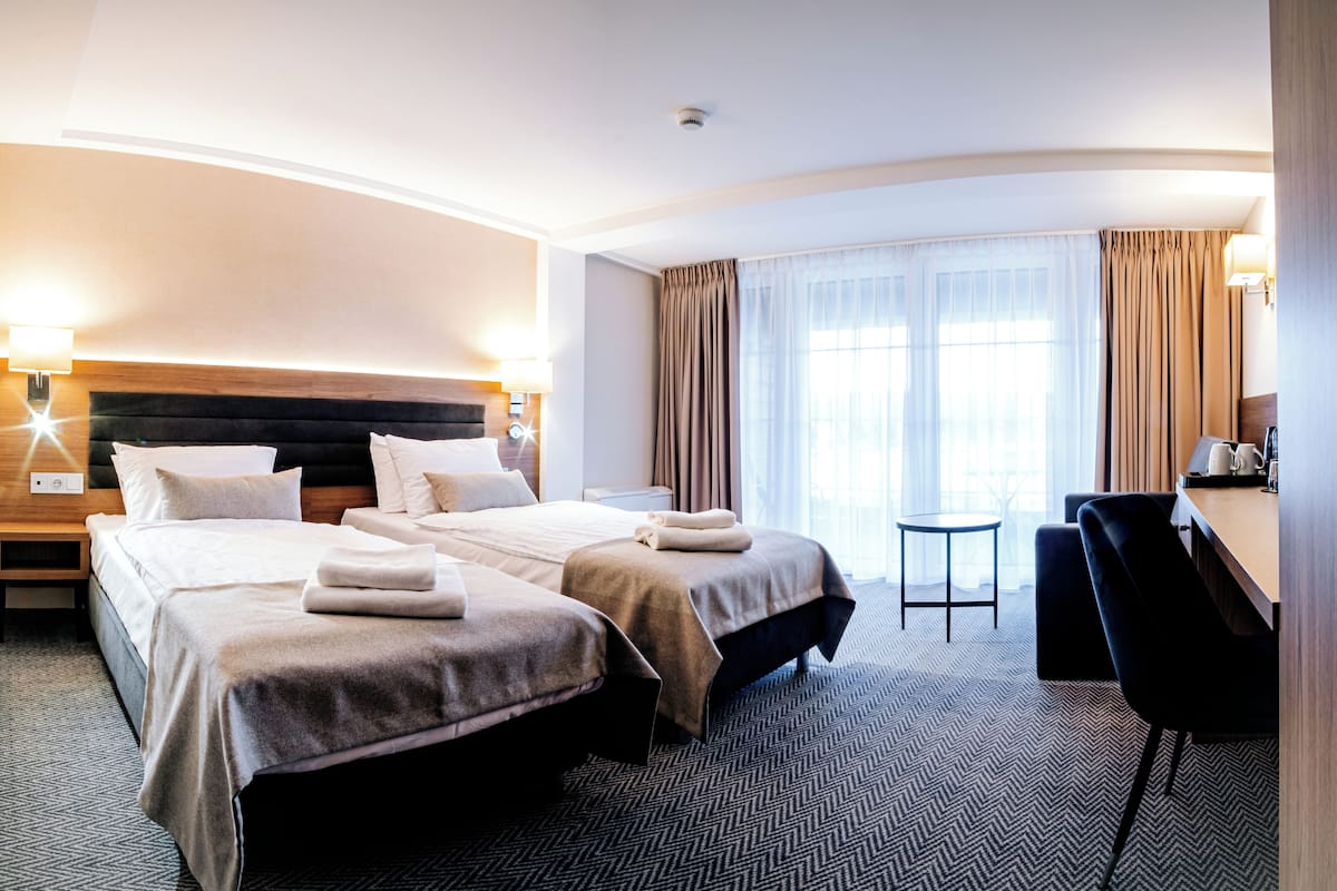 Une chambre d’hôtel avec deux lits, un bureau, une chaise, une petite table et une grande fenêtre avec des rideaux.