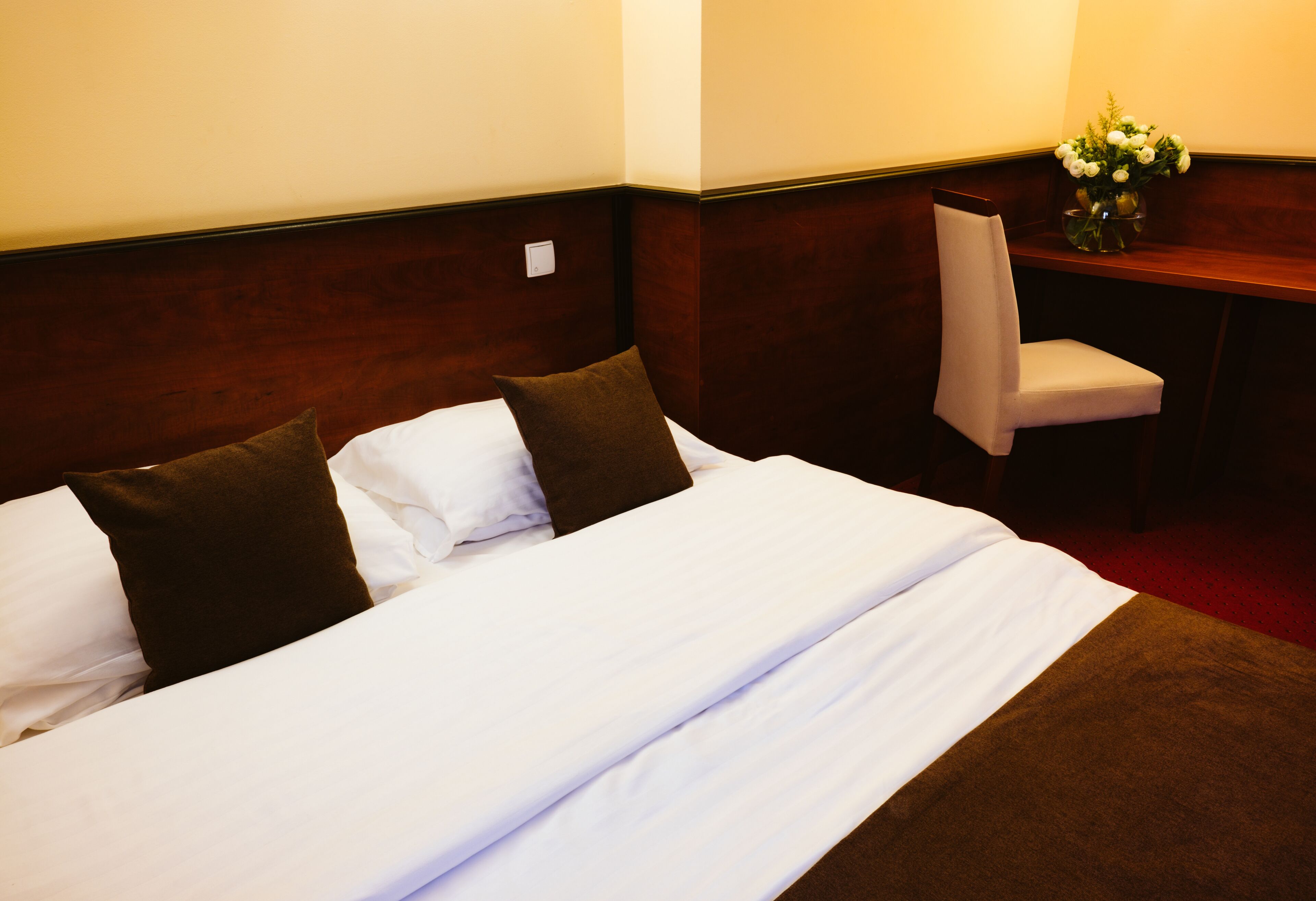 A hotel room with a neatly made bed, a chair, and a vase with flowers on a wooden table.