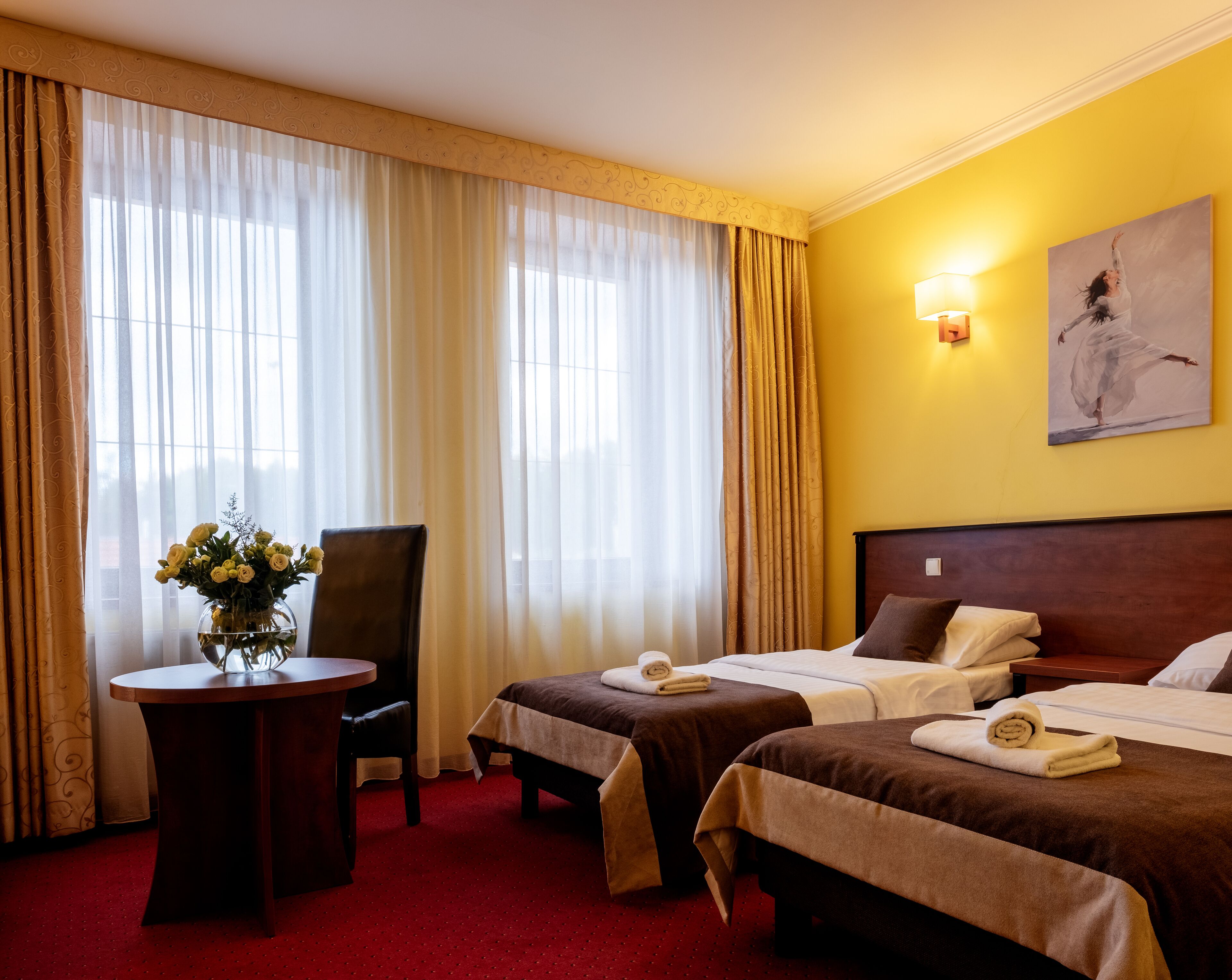 Ein Hotelzimmer mit zwei Betten, einem Schreibtisch mit Stuhl, einer Blumenvase und einem Gemälde an der Wand.